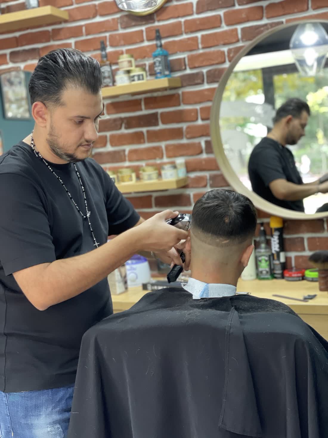 Frizerul lucrează cu mașina un fade pe ceafa unui client, în fața peretelui de cărămidă și a oglinzii rotunde de lemn