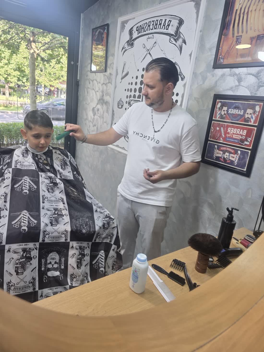Frizerul aranjează cu pieptănul tunsoarea unui băiat îmbrăcat în pelerina cu benzi desenate