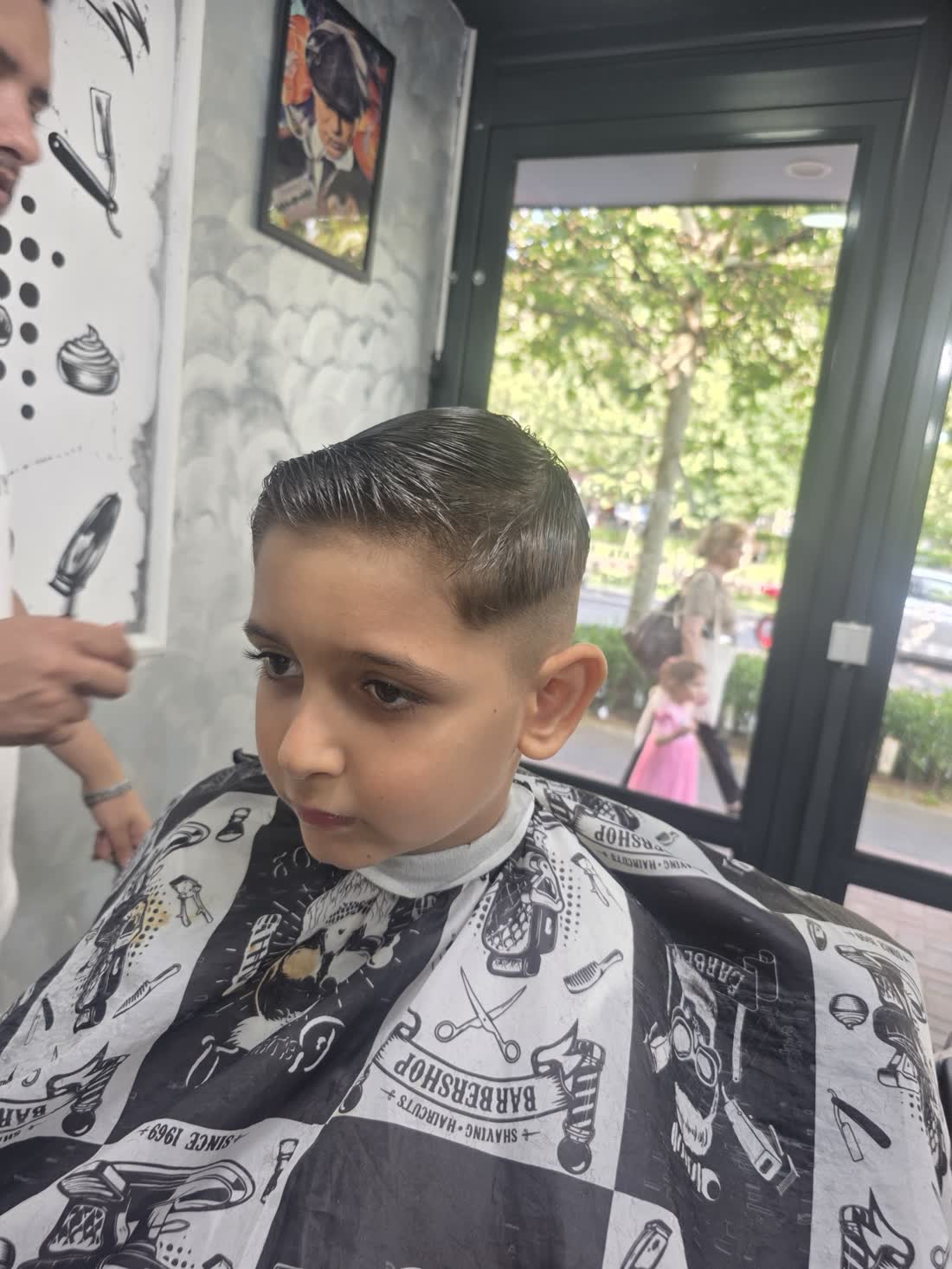Băiat tuns, în pelerina alb-negru, lângă geamul spre copacii de pe stradă